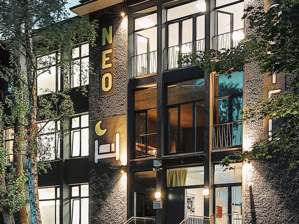 Beleuchtete Fassade des NEOHOSTEL mit Schriftzug und Eingangstreppe in der Abenddämmerung